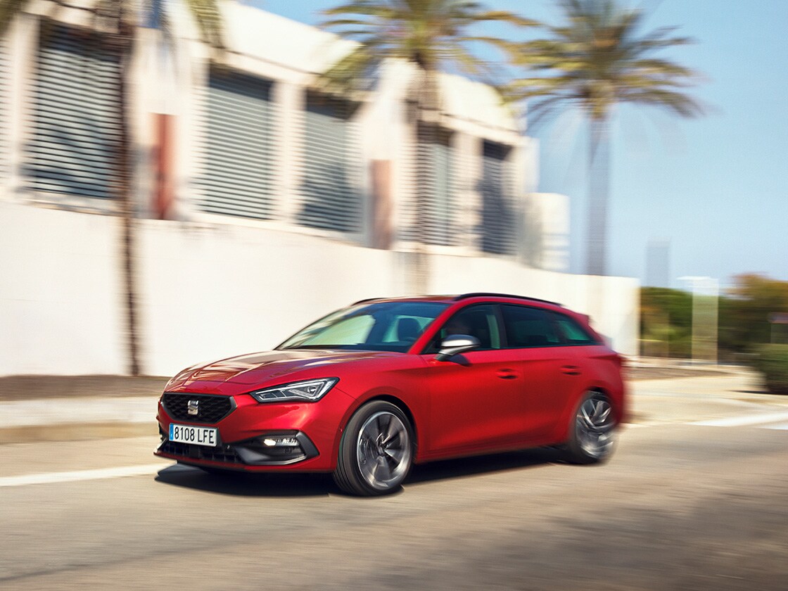 Una SEAT Leon Sportstourer rossa beneficia del programma di assistenza e manutenzione SEAT, mentre è in movimento in una zona residenziale con una casa e delle palme.